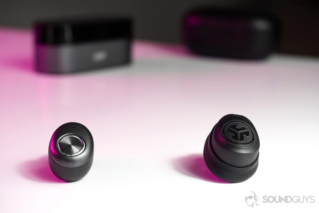 A picture of the JLab JBuds Air true wireless workout earbuds (black, right) next to the Monoprice true wireless earbuds (silver, left) with their respective charging cases out of focus in the background.