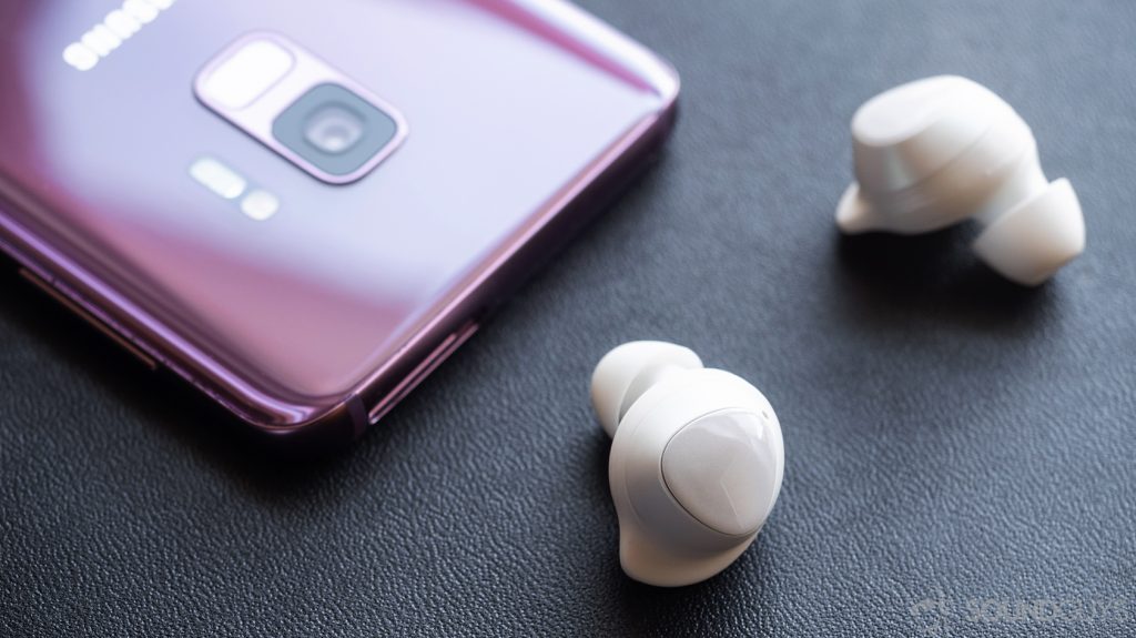 An angled image of the Samsung Galaxy Buds in front of a Samsung Galaxy S9 in Lilac.