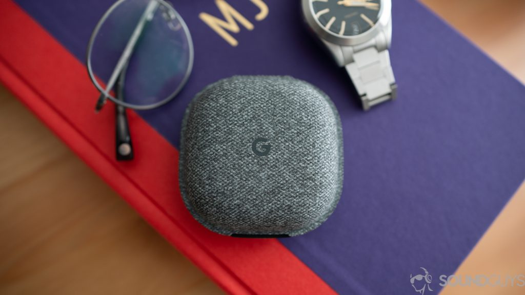Shot of the Pixel Buds charging case on a purple book next to a watch and glasses. 