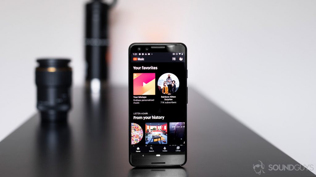 An image of a Google Pixel 3 with the YouTube Music Premium app open to the home screen. The phone is standing vertically on a black table with a lens and water bottle in the background.