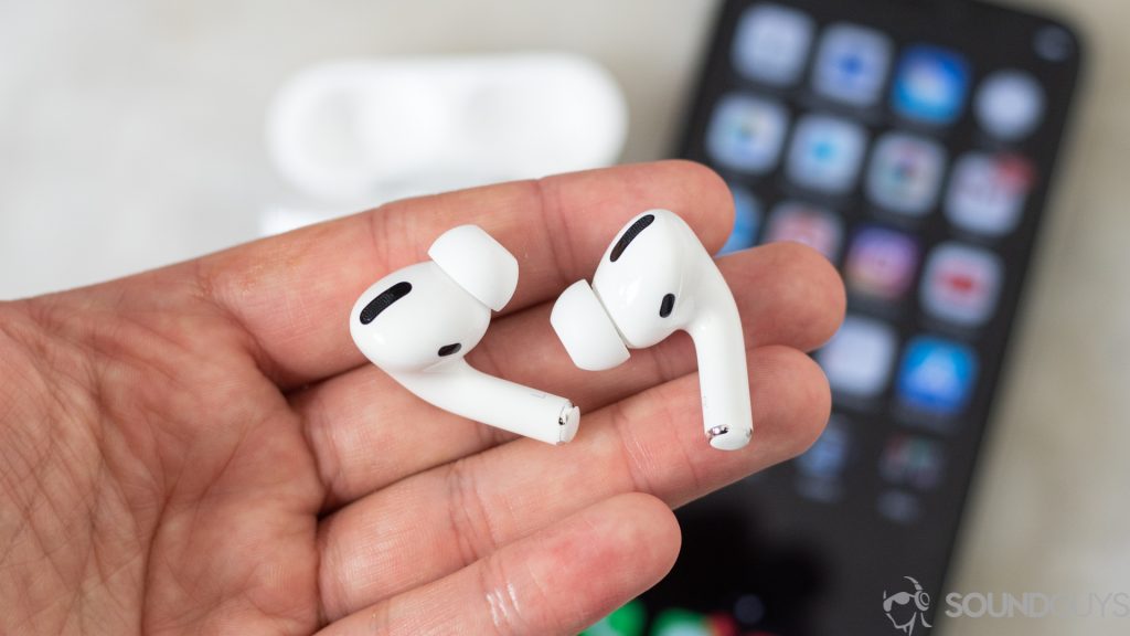 A picture of the Apple AirPods Pro in a man's left hand (foreground) with an iPhone and the AirPods Pro wireless charging case in the background; they are IPX4-rated and can be used as running earbuds..