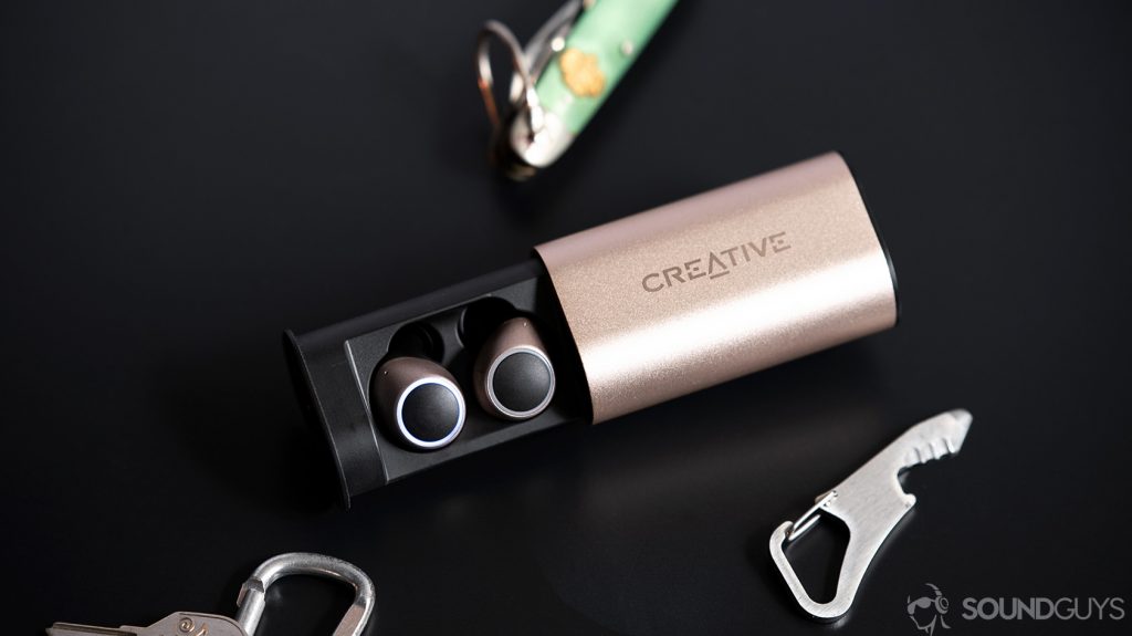 An image of the Creative Outlier Air earbuds in the case surrounded by an army knife, bottle opener, and carabiner on a black surface.