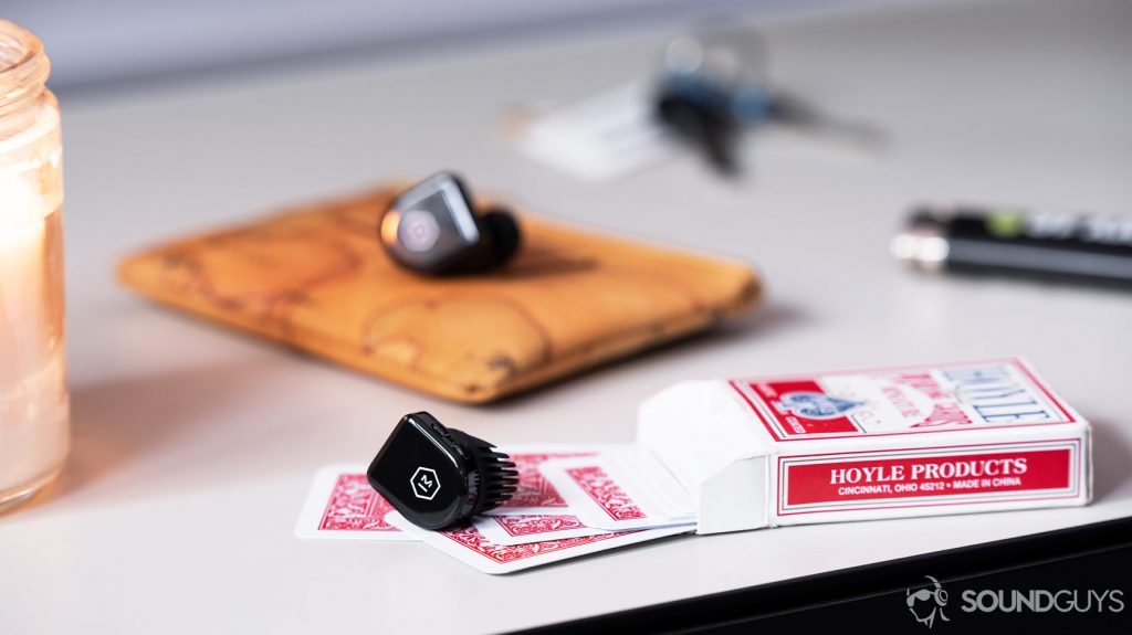 An image of the Master & Dynamic MW07 Go true wireless earbuds on red, miniature playing cards with a candle, keys, lighter, and coin pouch in the background.
