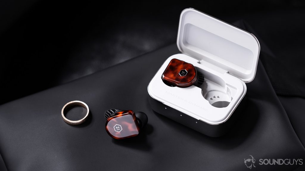 A picture of the Master & Dynamic MW07 Plus noise canceling true wireless earbuds with on earbud in the case and the other outside of it on a leather surface next to a gold ring.