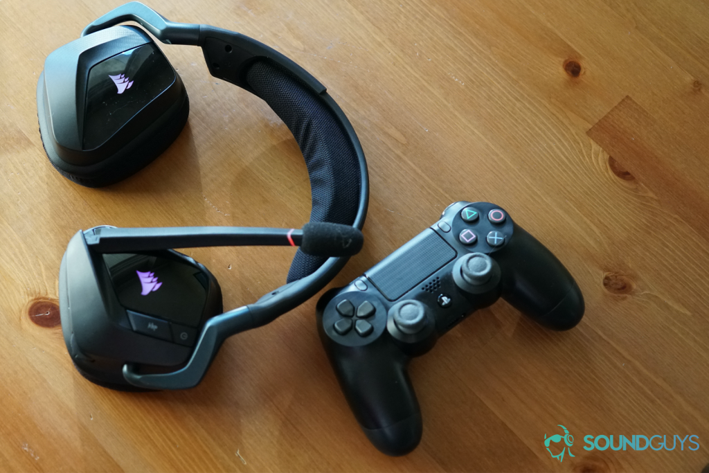 The Corsair Void RGB Elite Wireless gaming headset sites on a wooden table next to a Dualshock controller for the Sony Playstation 4