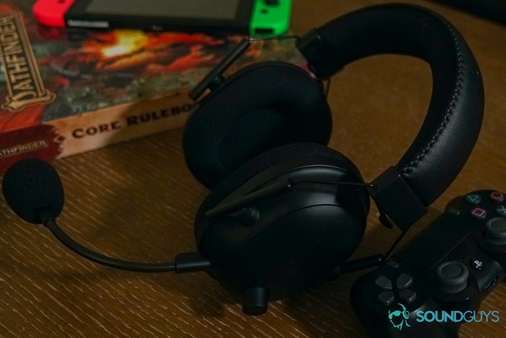 The Razer BlackShark V2 Pro sits on a wooden table next to a PlayStation Dualshock 4 controller, in front of a Nintendo Switch laying on a Pathfinder Rulebook.