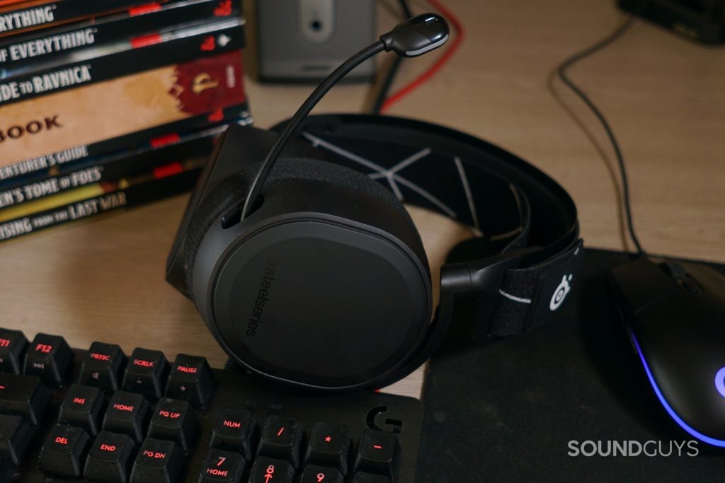 The SteelSeries Arctis 9 lays on a desk near a Logitech G4313 Carbon mechanical gaming keyboard, a Logitech gaming mouse, a Bose companion speaker, and various rule books for Dungeons and Dragons and Pathfinder.
