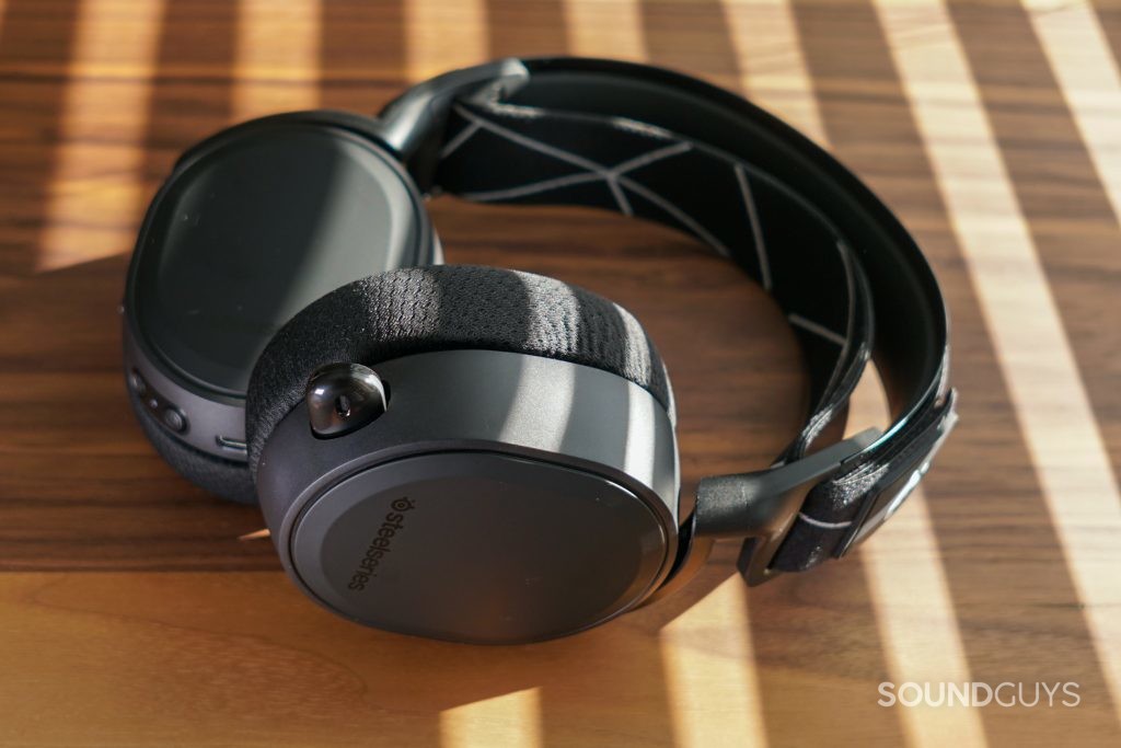 The SteelSeries Arctis 9 gaming headset lays on a wooden table with sunlight streaming through blinds.