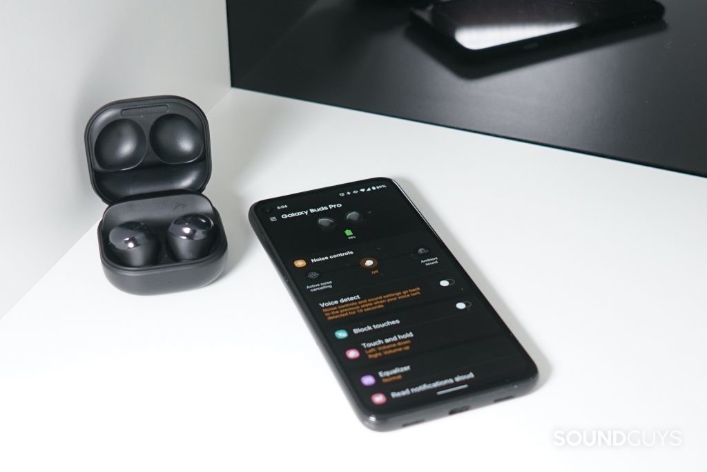 The Samsung Galaxy Buds Pro sit on a shelf next to a Google Pixel 4a with the Samsung Wearables app open on it.