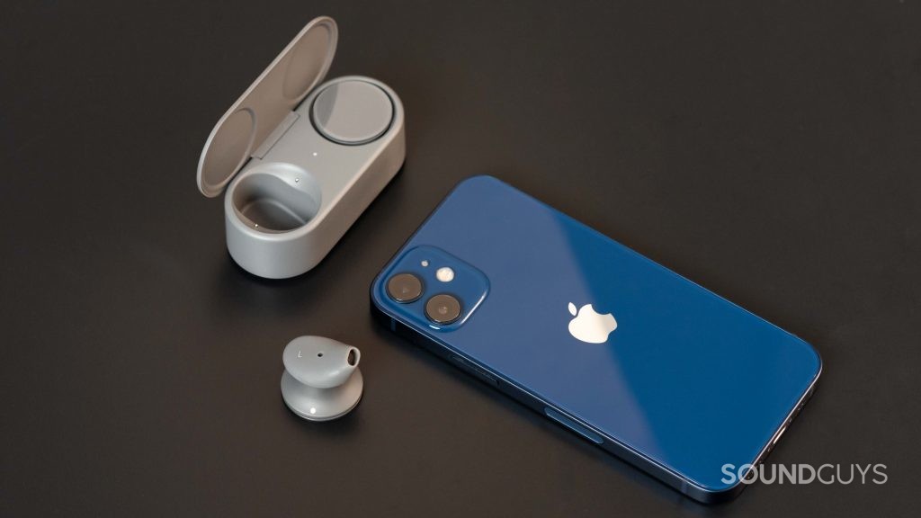 The Microsoft Surface Earbuds with one bud in the case and one outside of it next to an iPhone 12 Mini in blue.