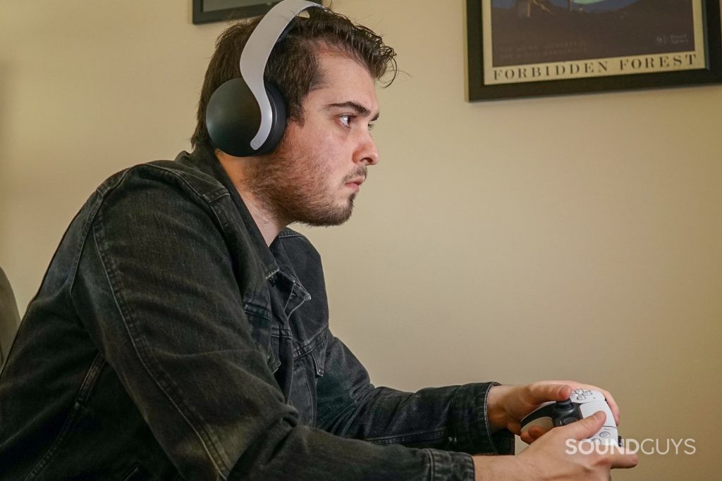 A man sits on a couch playing on a PlayStation 5 while wearing the Sony Pulse 3D Wireless Headset and holding a PlayStation DualSense controller.