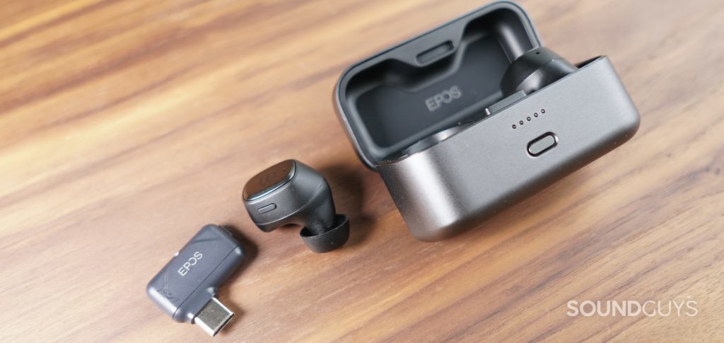The EPOS GTW 270 Hybrid true wireless earbuds sit on a table with one earbud out of the case, and the USB-C dongle laying next to it.