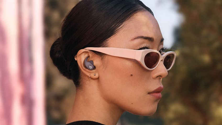 A woman wearing sunglasses shown looking to the right wearing Beats Fit Pro earbuds in grey.