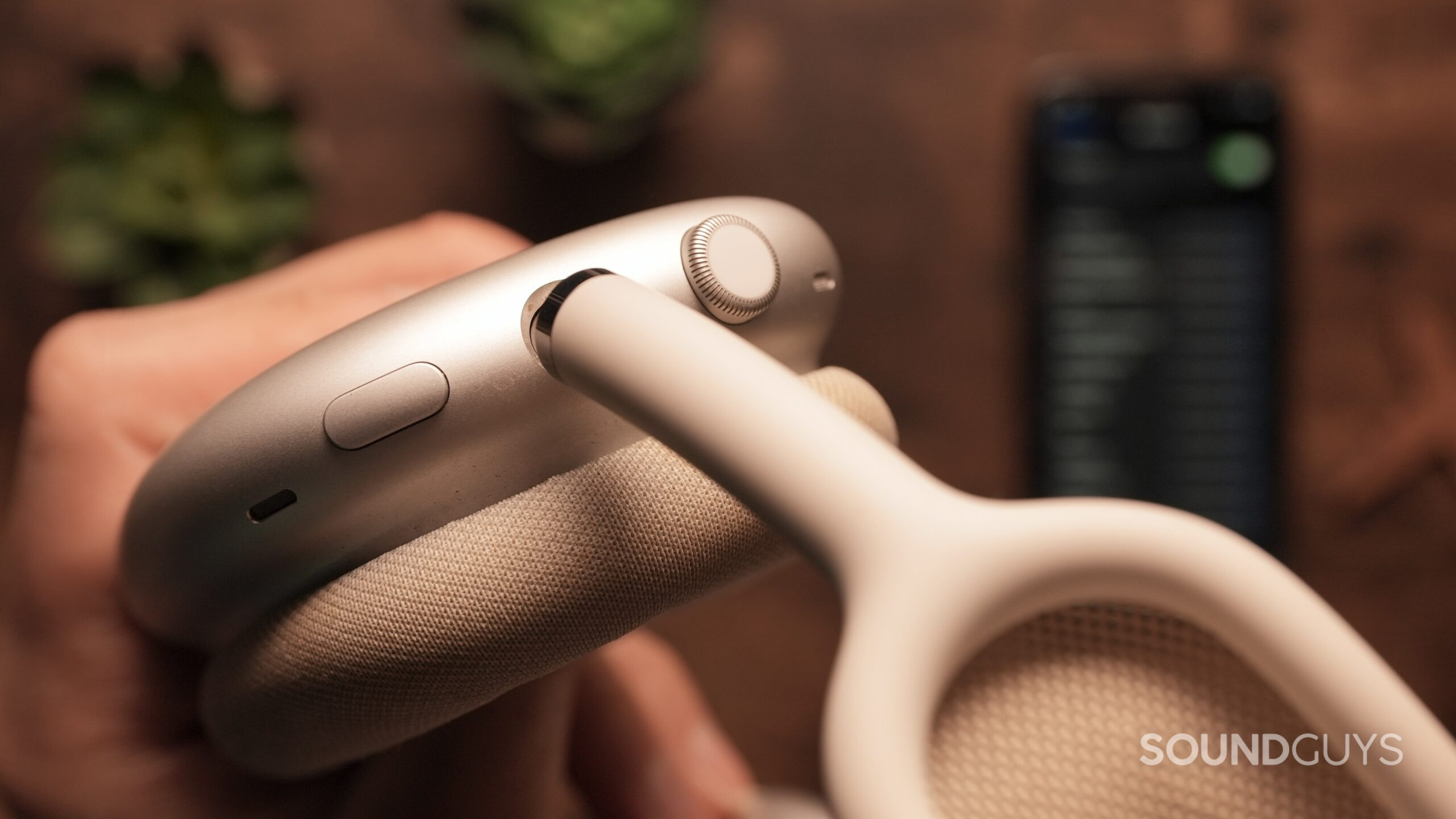 A hand holds an AirPods Max, showing the digital crown and noise control buttons.