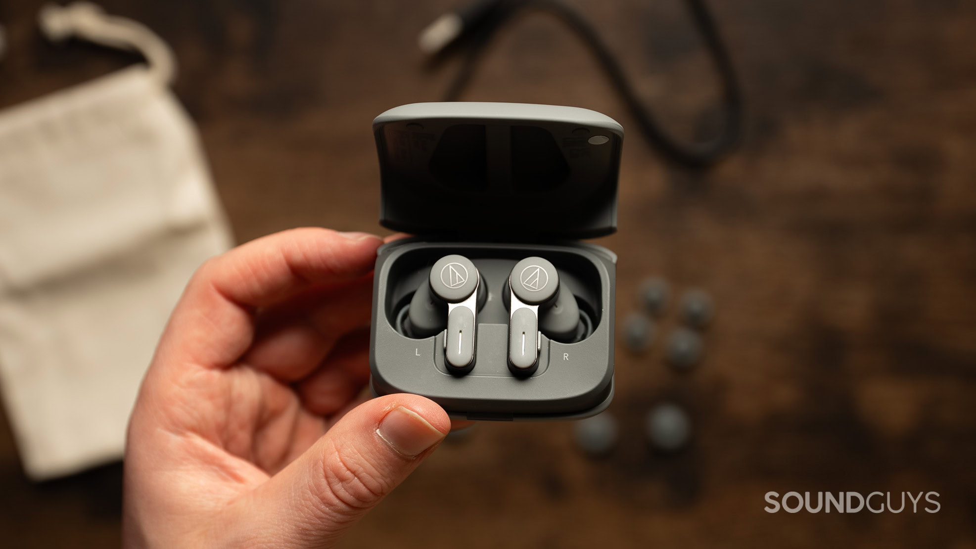 A hand holds the Audio-Technica ATH-TWX7 earbuds inside their charging case over a wood table. 