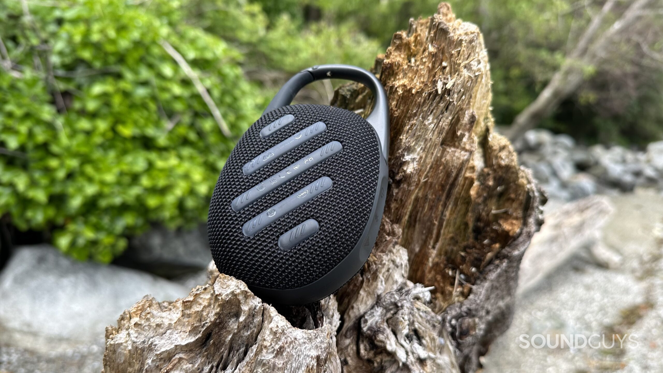 A JBL Clip 5 resting on a piece of beach driftwood, showing the silicon padding backside.