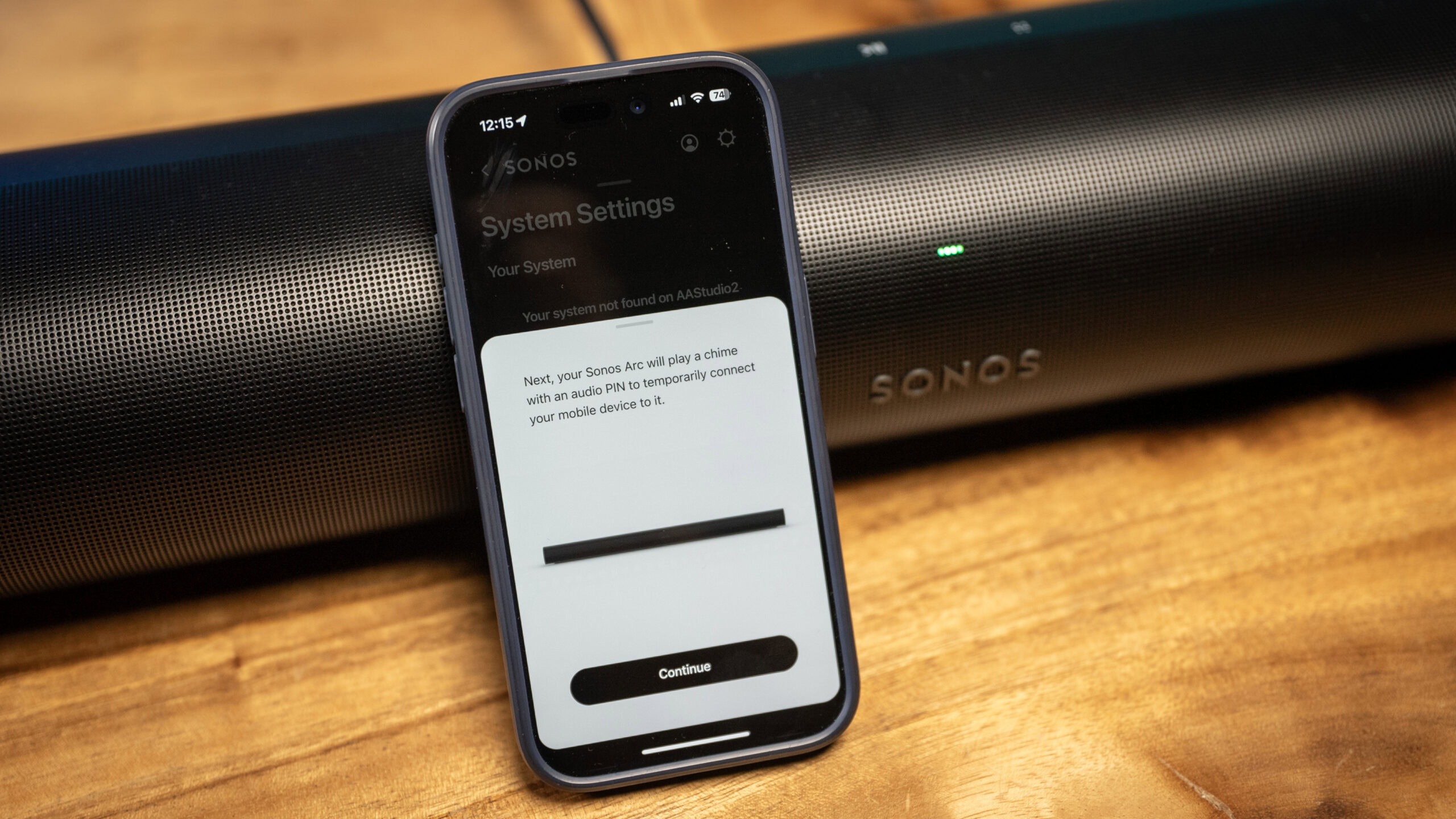 An iPhone leaning against the Sonos Arc soundbar on a wooden table. The phone shows the connection process for the Sonos Arc via the Sonos app.