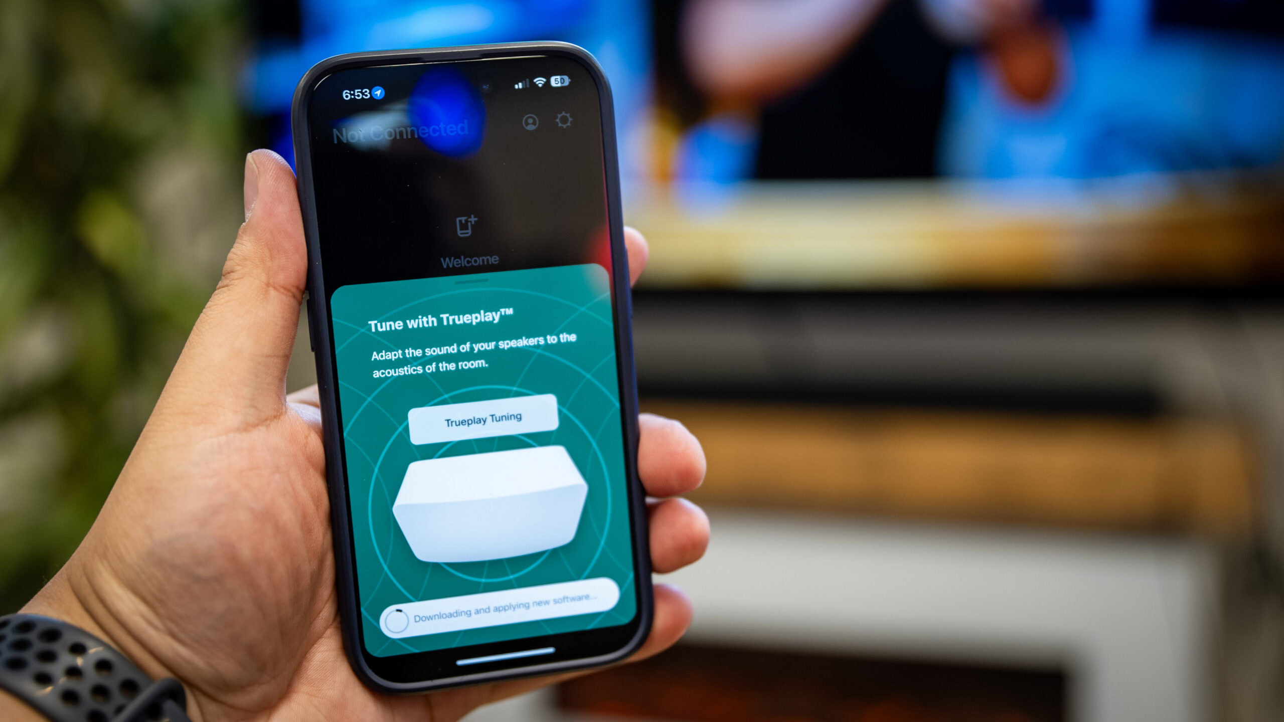 A man holding an iPhone with the Sonos app open showing the Trueplay tuning feature for the Sonos Arc soundbar. TV and soundbar are in the background out of focus.