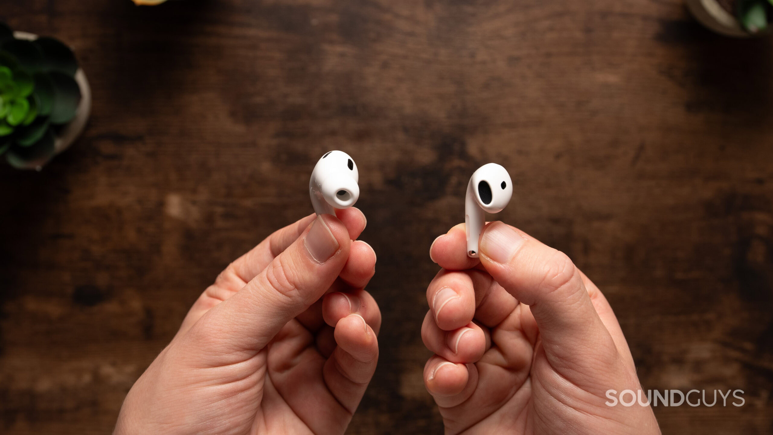 One hand holds an AirPods 4 earbud without an ear tip while another hand holds an AirPods Pro 2 earbud with an ear tip.