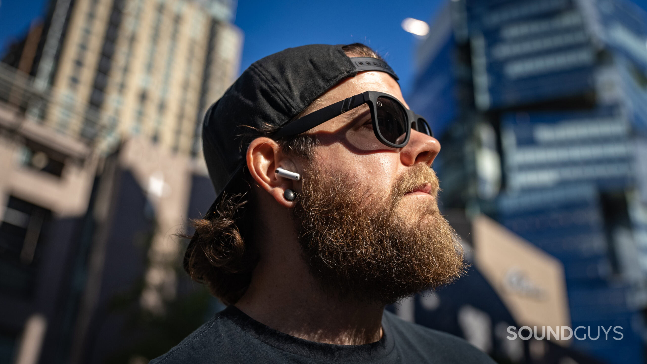 A man wearing an AirPods 4 earbud outside in the sunlight surrounded by buildings.