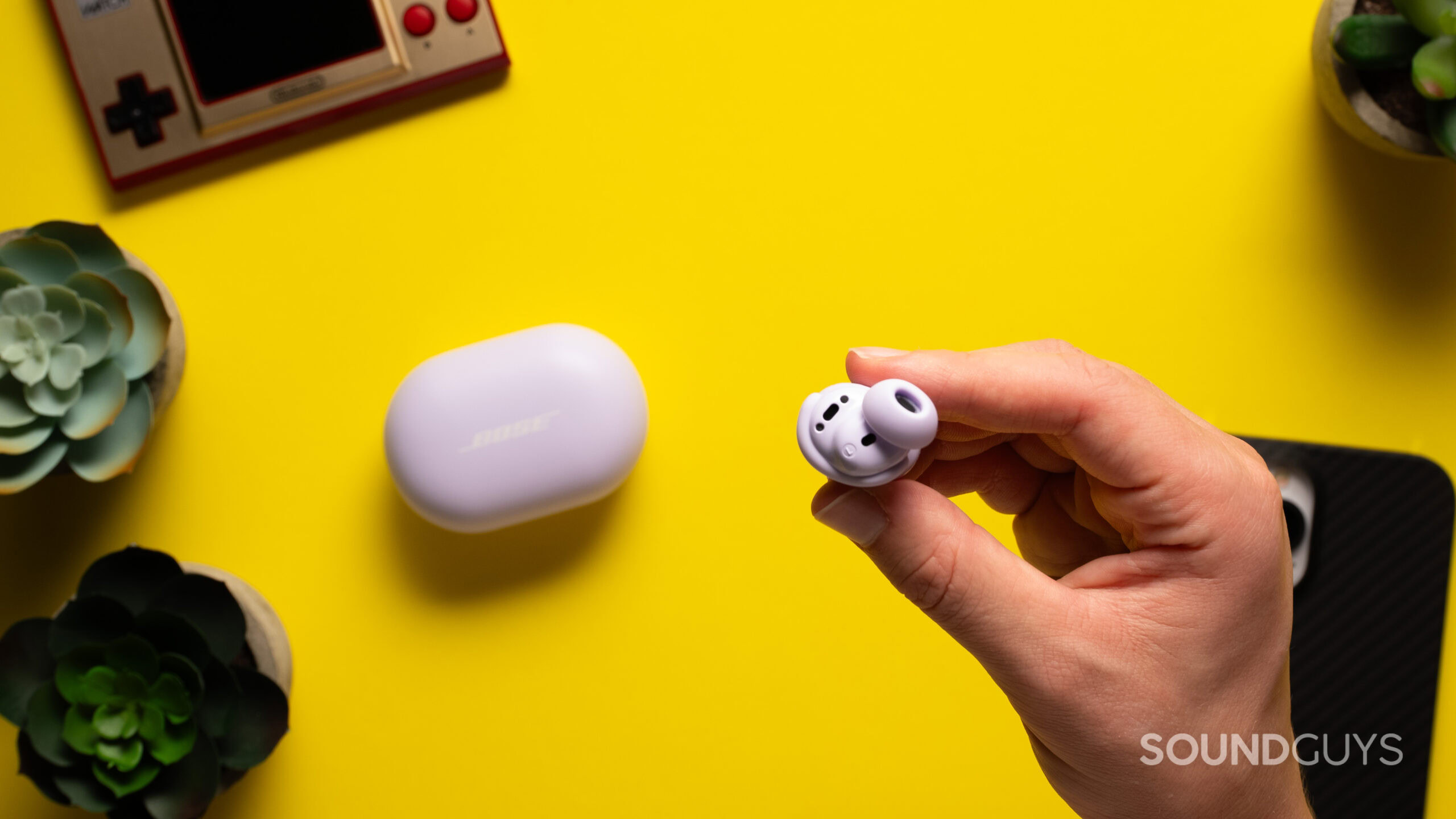 A hand holds a Bose QuietComfort Earbuds bud above a table with the charging case.