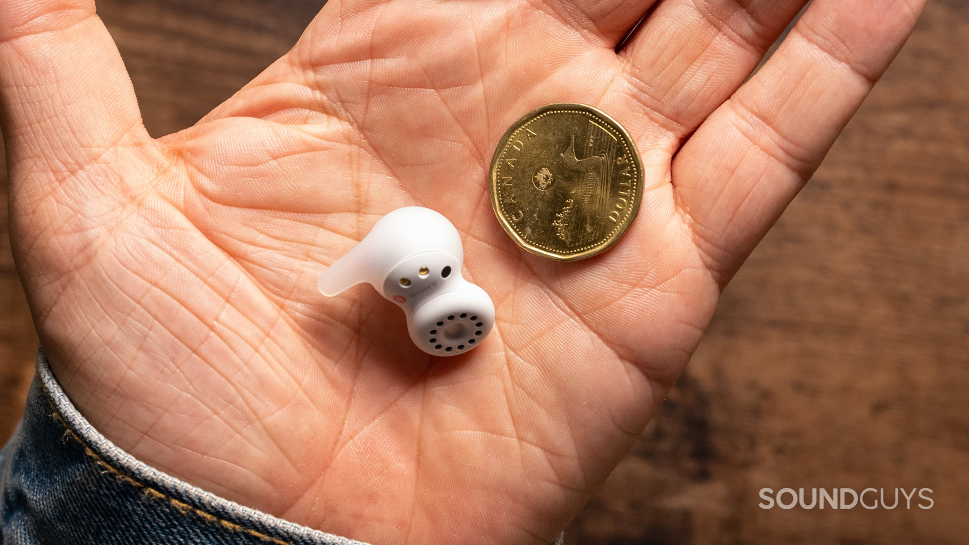 A photo of the Sony LinkBuds Open in a man's hand, next to a Canadian $1 coin.