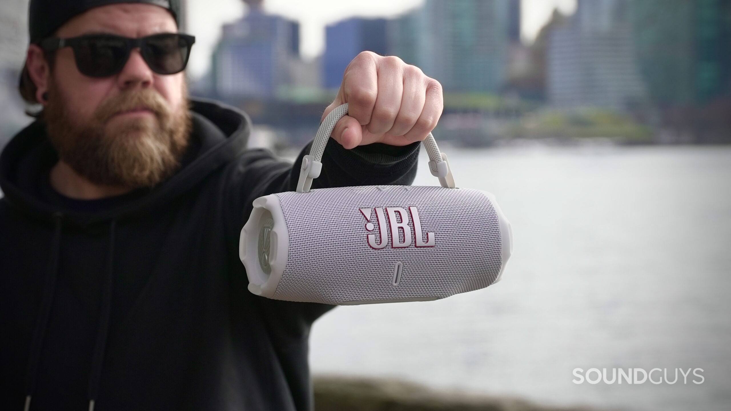 A man holds the JBL Flip Charge 6 speaker by its handle with the city in the back ground.