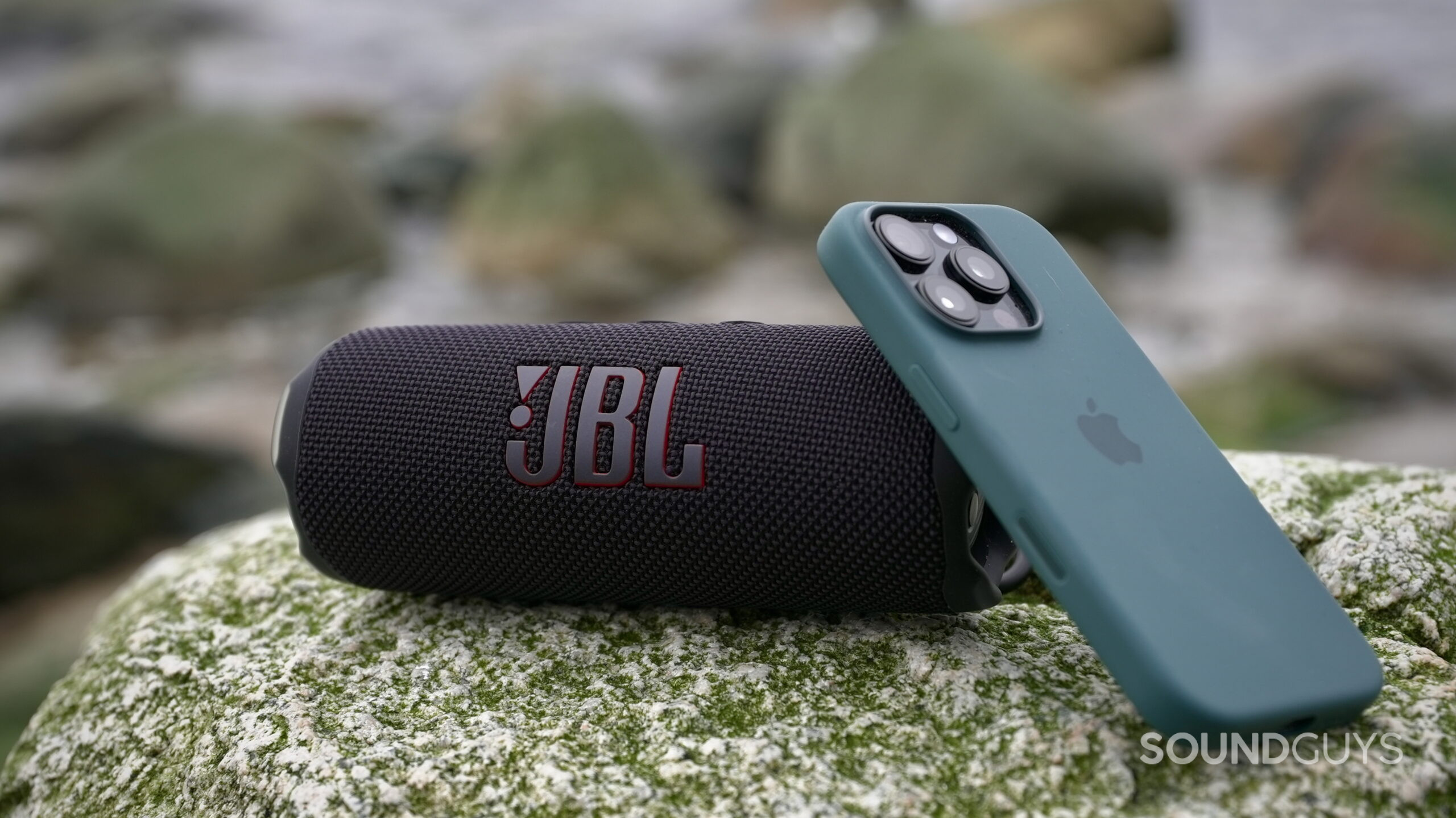 A JBL Flip 7 placed on as rock, and an iPhone 16 Pro leaving up against it.